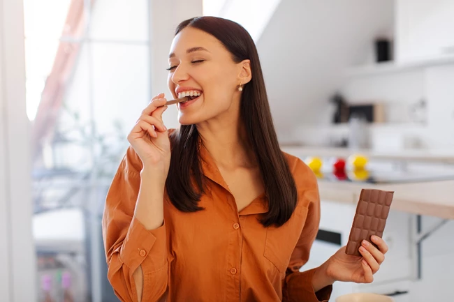 Happy woman enjoying taste of chocolate bar, sitting in kitchen at home