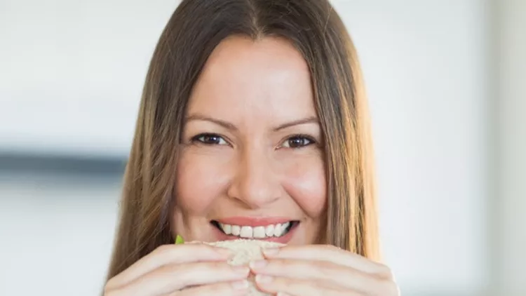 portrait-of-a-smiling-woman-eating-sandwich-in-kitchen-picture-id493203561