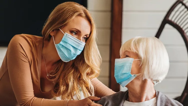 senior-woman-in-medical-mask-with-social-worker-visiting-her-at-home-picture-id1261423020 (1)